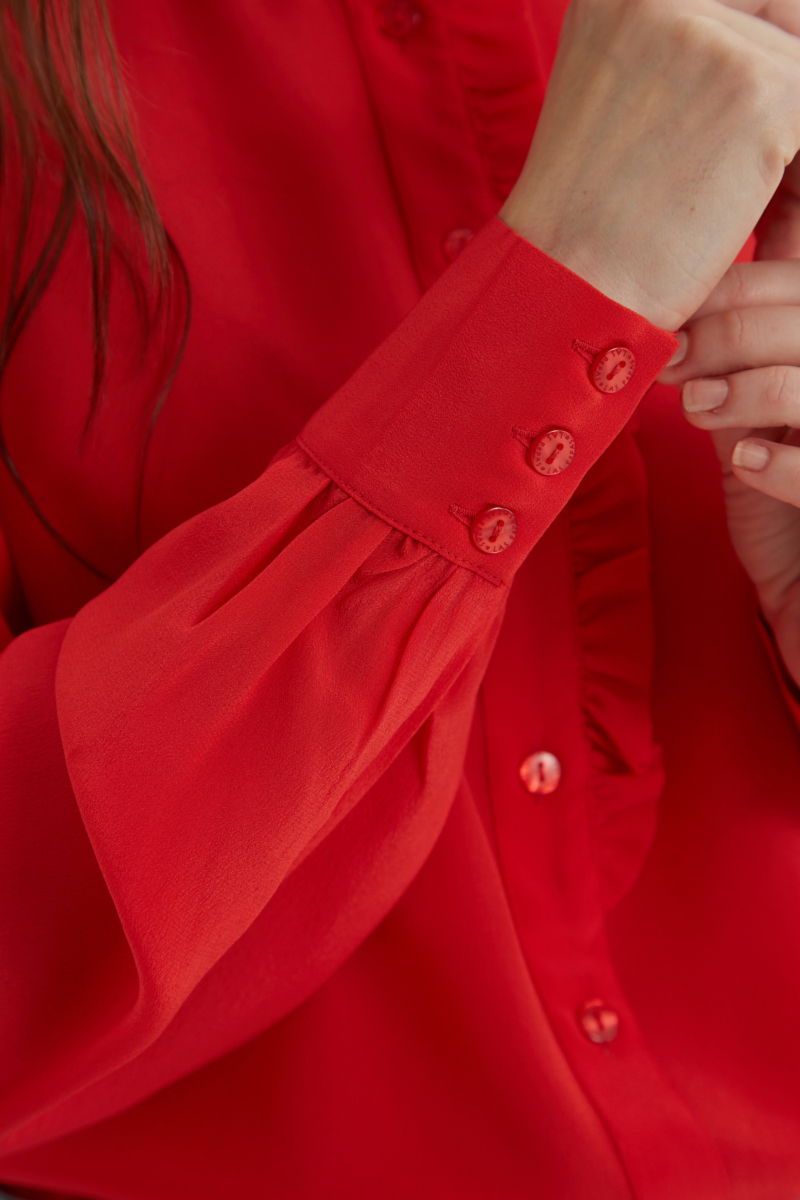 Camisa roja de seda recta y larga a la cadera con cuello mao y volados en el cuello y la cartera, con frunces en los hombros y puños y botones a tono. 