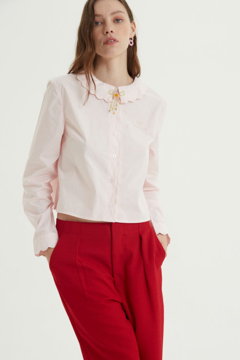 Camisa rosa de poplin de largo hasta la cadera con terminación en forma de onditas en cuello, puños y cartera y botones a tono en forma de flor. 