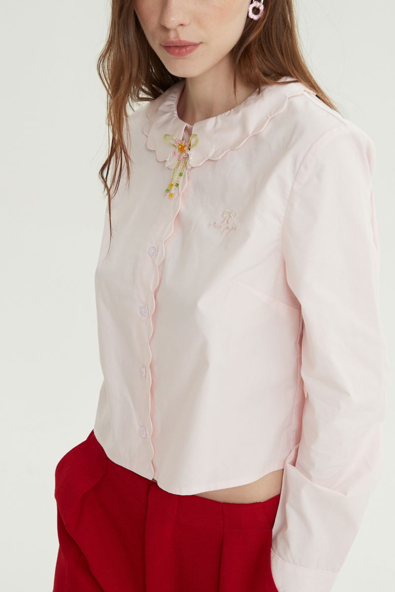 Camisa rosa de poplin de largo hasta la cadera con terminación en forma de onditas en cuello, puños y cartera y botones a tono en forma de flor. 