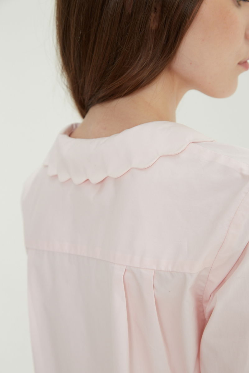 Camisa rosa de poplin de largo hasta la cadera con terminación en forma de onditas en cuello, puños y cartera y botones a tono en forma de flor. 
