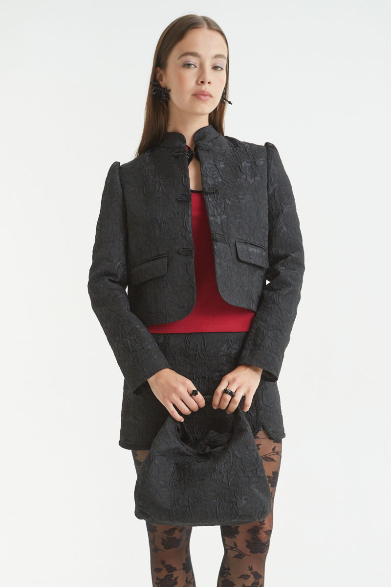Saco negro de brocato con textura de rosas y alamares de pasamanería, mangas princesas y cuello mao, con bolsillos con solapa delanteros y largo a la cadera. 