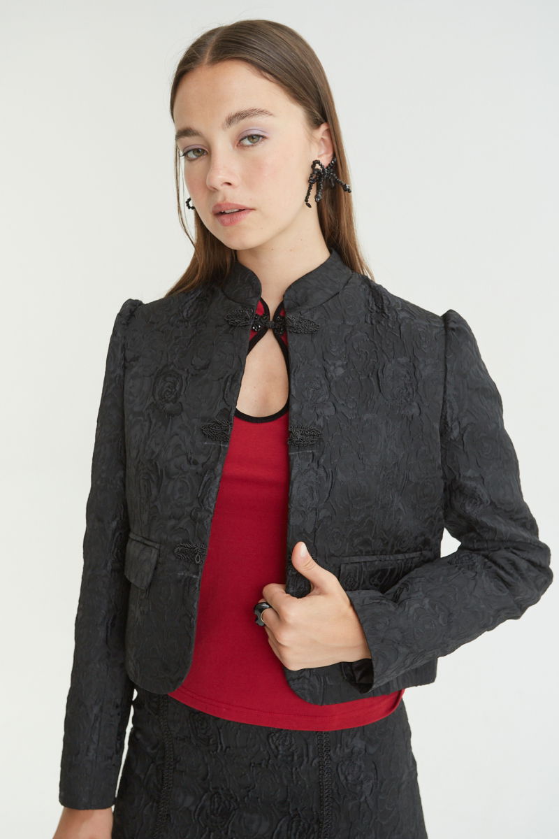 Saco negro de brocato con textura de rosas y alamares de pasamanería, mangas princesas y cuello mao, con bolsillos con solapa delanteros y largo a la cadera. 