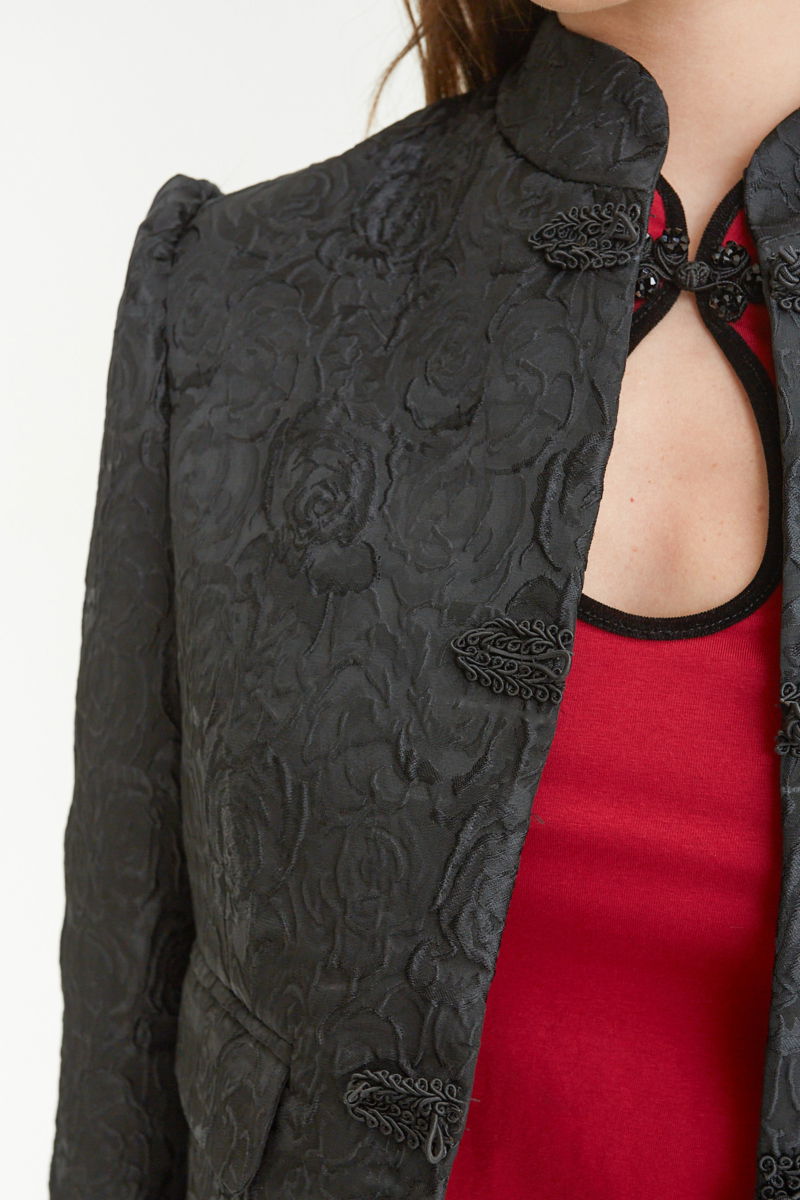 Saco negro de brocato con textura de rosas y alamares de pasamanería, mangas princesas y cuello mao, con bolsillos con solapa delanteros y largo a la cadera. 