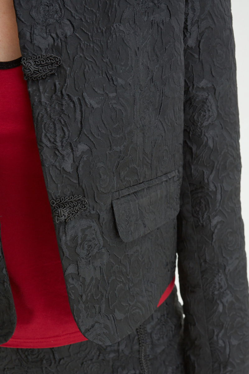 Saco negro de brocato con textura de rosas y alamares de pasamanería, mangas princesas y cuello mao, con bolsillos con solapa delanteros y largo a la cadera. 