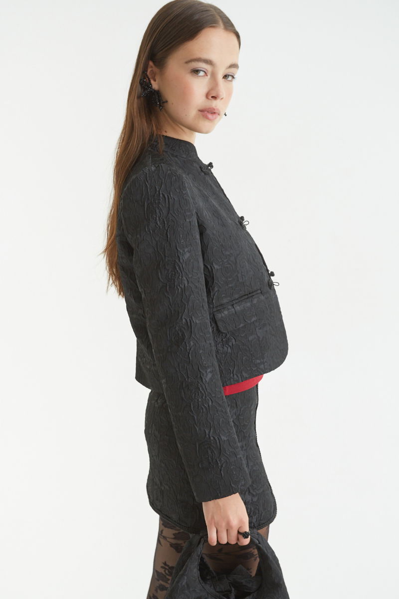 Saco negro de brocato con textura de rosas y alamares de pasamanería, mangas princesas y cuello mao, con bolsillos con solapa delanteros y largo a la cadera. 