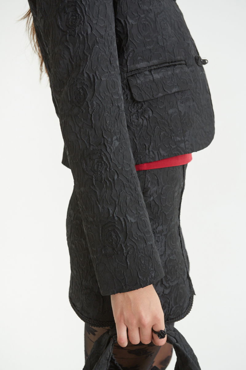 Saco negro de brocato con textura de rosas y alamares de pasamanería, mangas princesas y cuello mao, con bolsillos con solapa delanteros y largo a la cadera. 