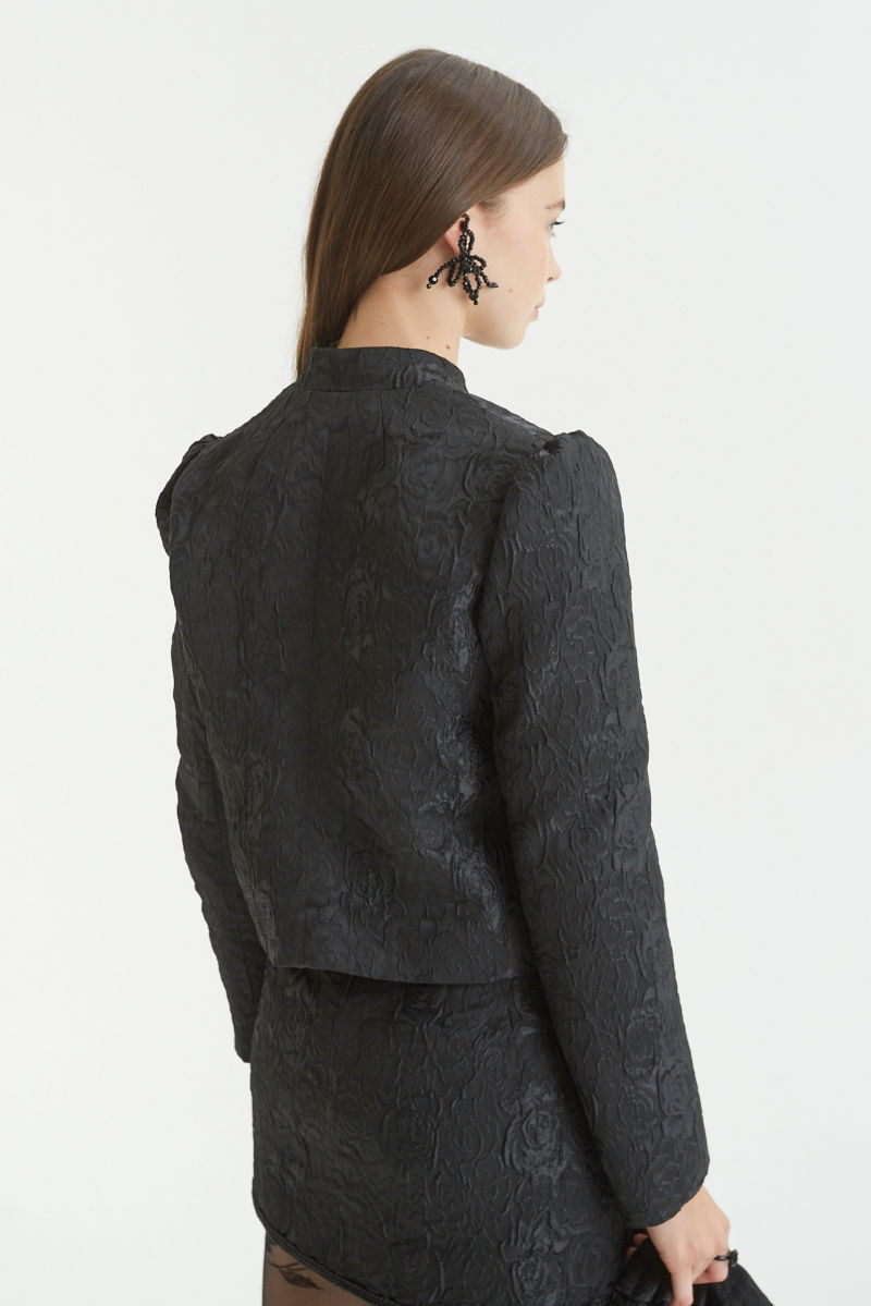 Saco negro de brocato con textura de rosas y alamares de pasamanería, mangas princesas y cuello mao, con bolsillos con solapa delanteros y largo a la cadera. 