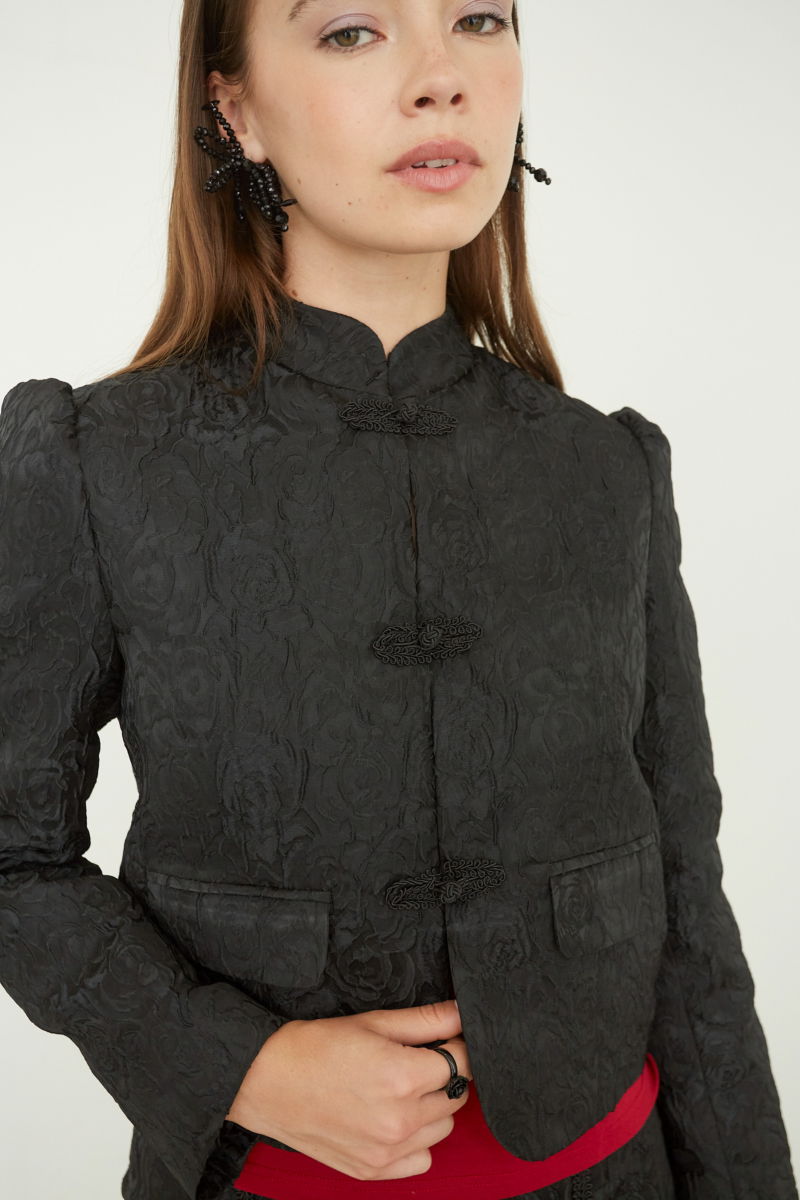 Saco negro de brocato con textura de rosas y alamares de pasamanería, mangas princesas y cuello mao, con bolsillos con solapa delanteros y largo a la cadera. 