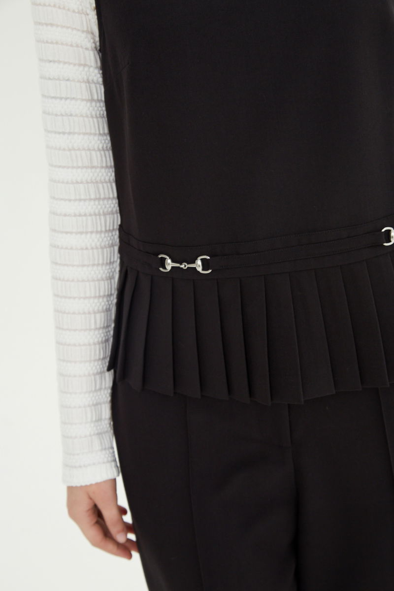 Blusa negra sastrera sin mangas con tablas en la cintura, herrajes metálicos y escote a la base del cuello, larga a la cadera. 