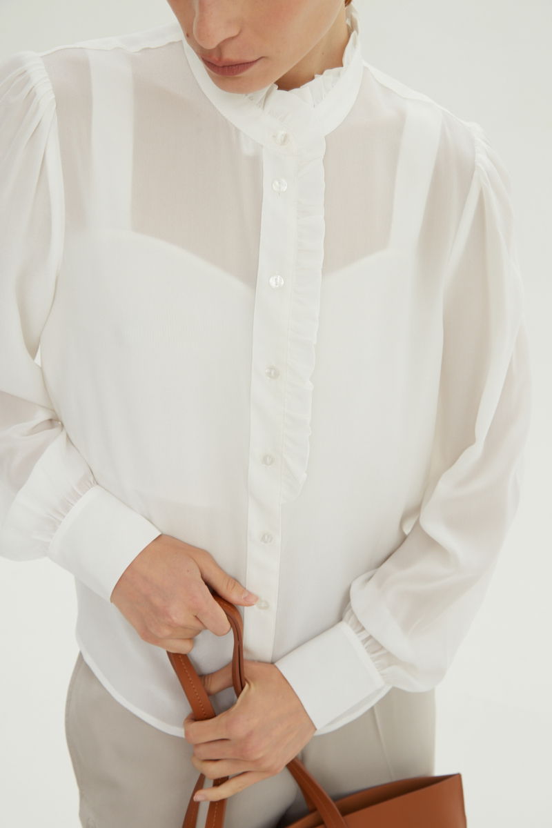 Camisa cruda de seda recta y larga a la cadera con cuello mao y volados en el cuello y la cartera, con frunces en los hombros y puños y botones a tono. 