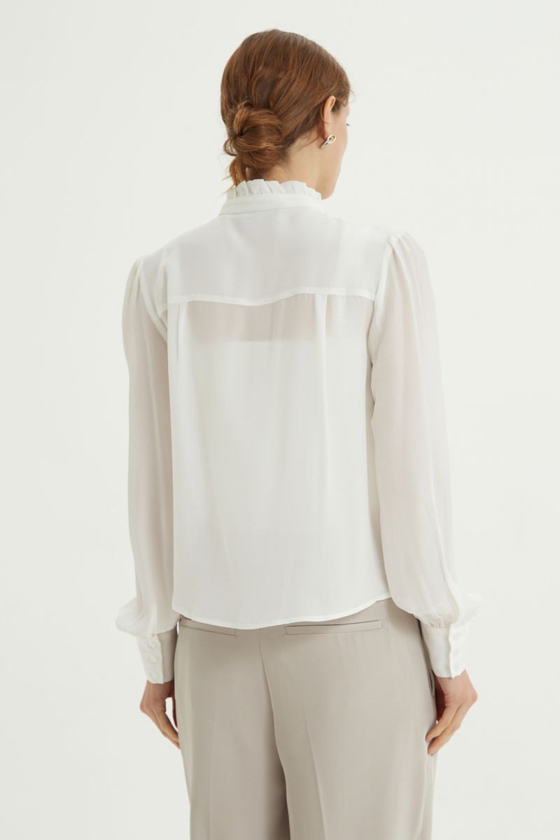 Camisa cruda de seda recta y larga a la cadera con cuello mao y volados en el cuello y la cartera, con frunces en los hombros y puños y botones a tono. 