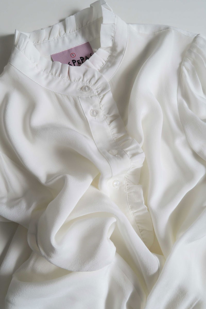 Camisa cruda de seda recta y larga a la cadera con cuello mao y volados en el cuello y la cartera, con frunces en los hombros y puños y botones a tono. 