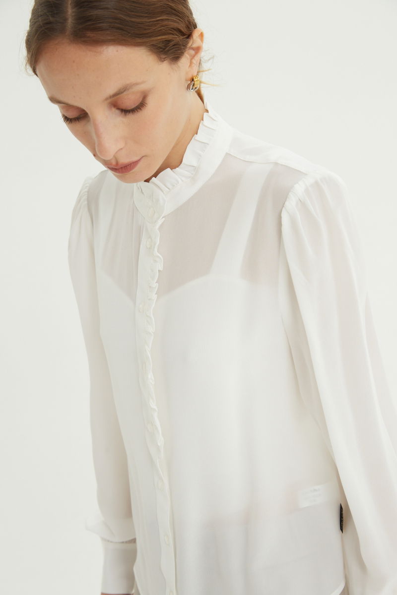 Camisa cruda de seda recta y larga a la cadera con cuello mao y volados en el cuello y la cartera, con frunces en los hombros y puños y botones a tono. 