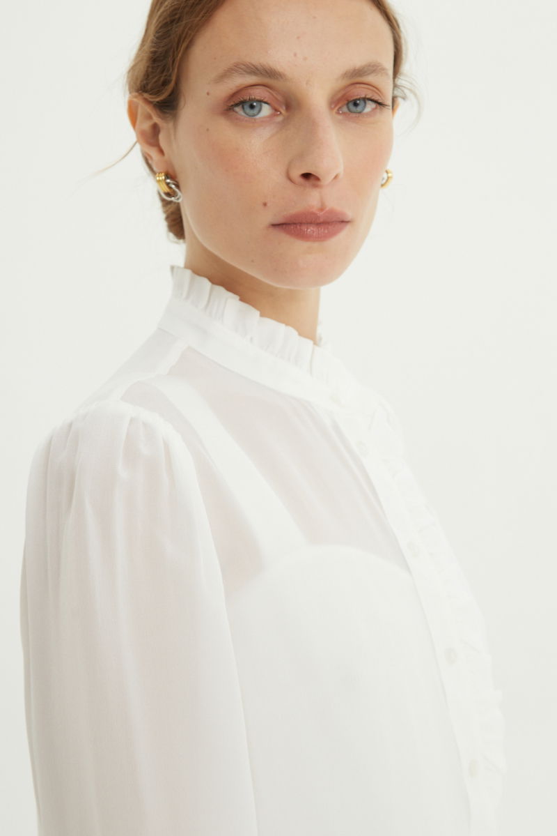 Camisa cruda de seda recta y larga a la cadera con cuello mao y volados en el cuello y la cartera, con frunces en los hombros y puños y botones a tono. 