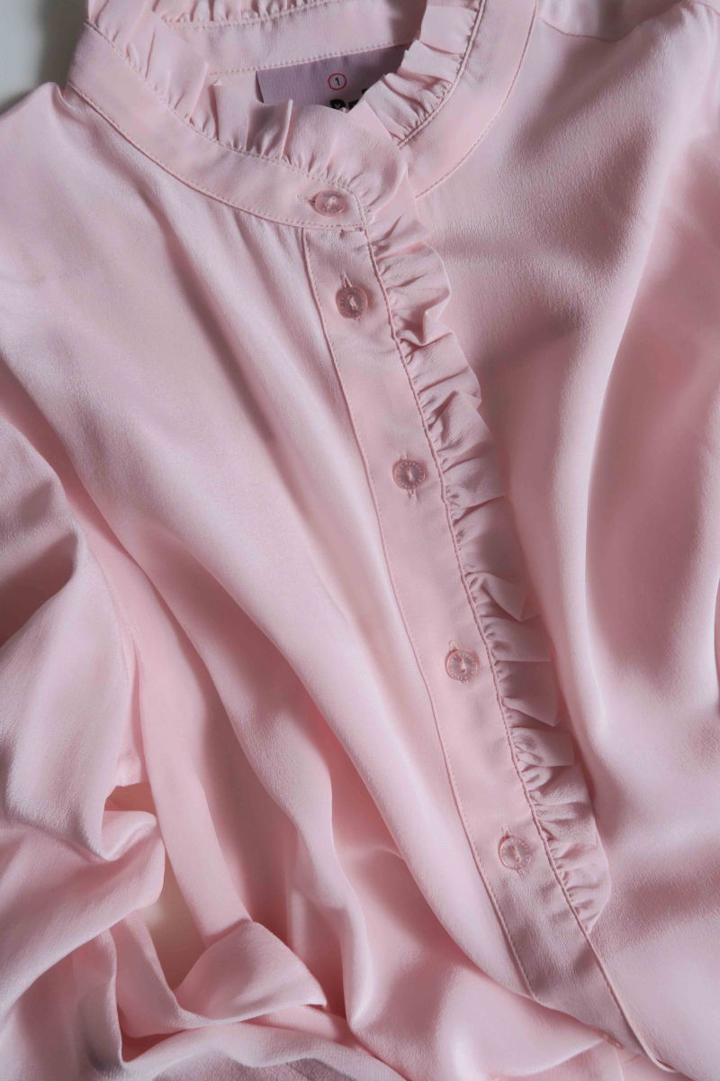 Camisa rosa de seda recta y larga a la cadera con cuello mao y volados en el cuello y la cartera, con frunces en los hombros y puños y botones a tono. 