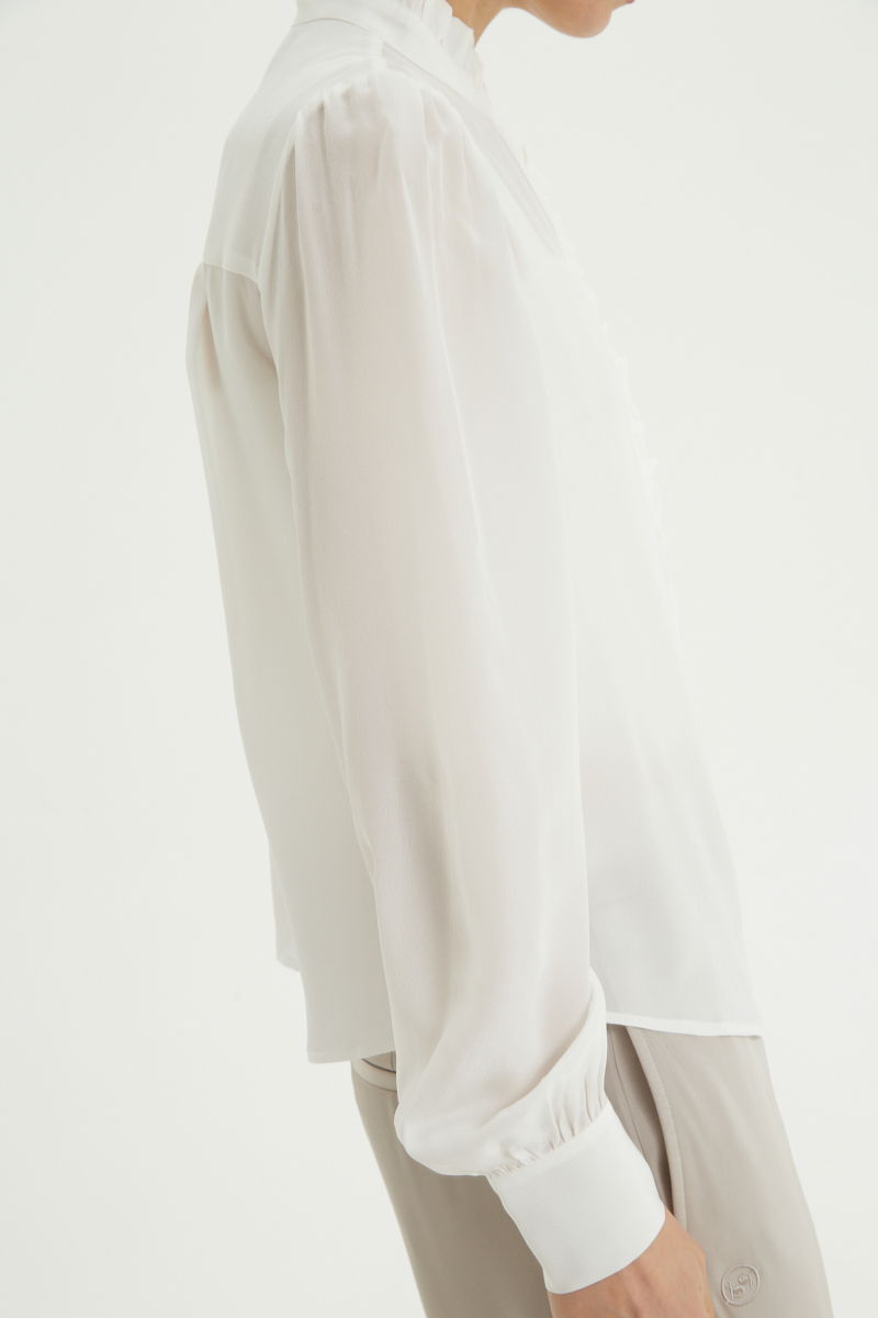 Camisa cruda de seda recta y larga a la cadera con cuello mao y volados en el cuello y la cartera, con frunces en los hombros y puños y botones a tono. 