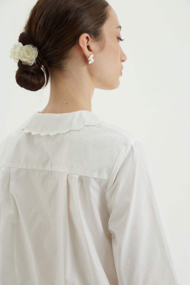 Camisa blanco de poplin de largo hasta la cadera con terminación en forma de onditas en cuello, puños y cartera y botones a tono en forma de flor. 