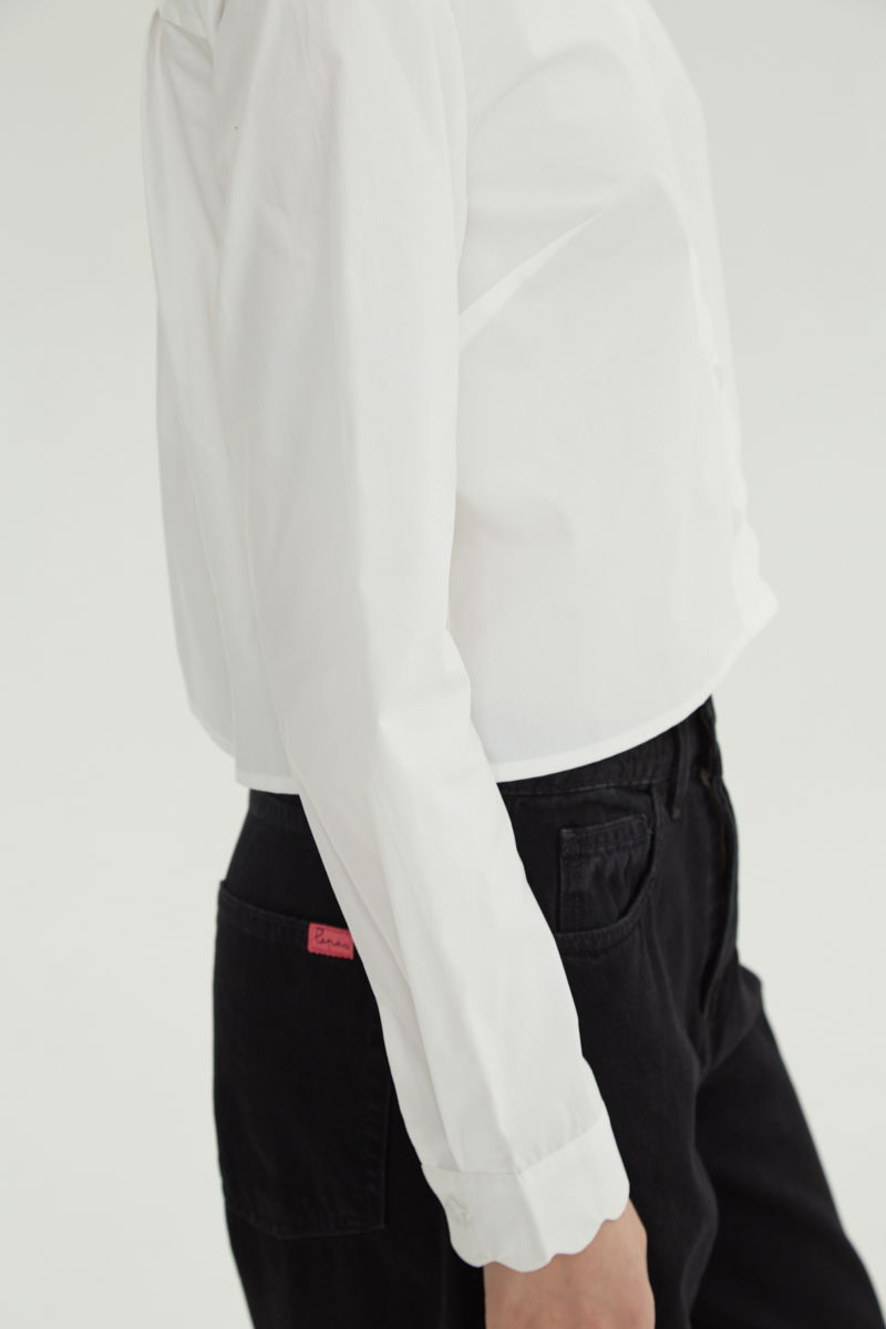 Camisa blanco de poplin de largo hasta la cadera con terminación en forma de onditas en cuello, puños y cartera y botones a tono en forma de flor. 