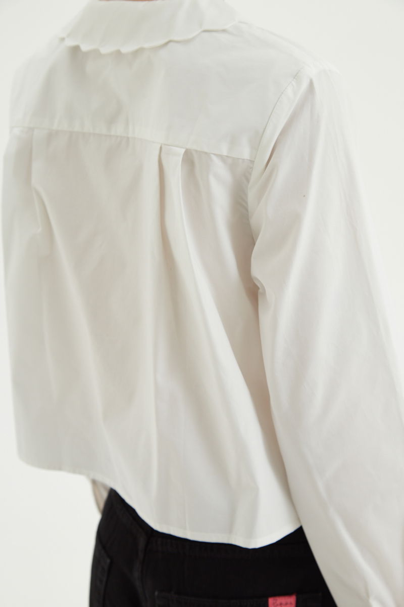 Camisa blanco de poplin de largo hasta la cadera con terminación en forma de onditas en cuello, puños y cartera y botones a tono en forma de flor. 