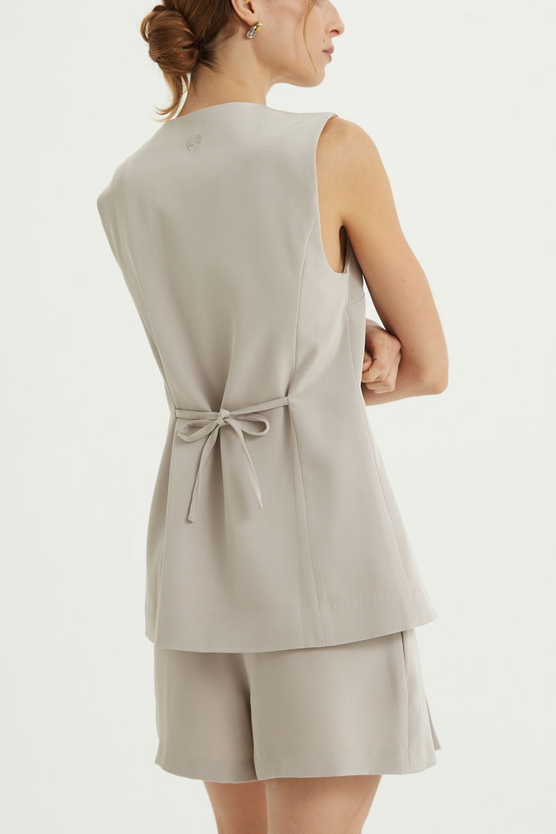 Chaleco sastrero beige con escote en V y largo a la cadera con botones a tono y lazo en la espalda. 