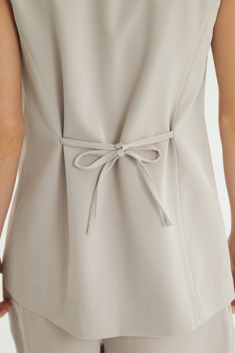 Chaleco sastrero beige con escote en V y largo a la cadera con botones a tono y lazo en la espalda. 