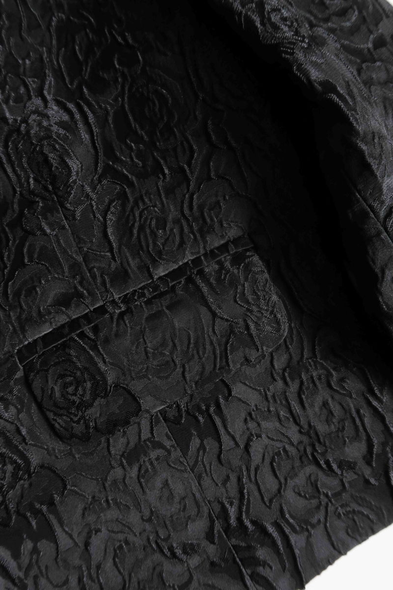 Saco negro de brocato con textura de rosas y alamares de pasamanería, mangas princesas y cuello mao, con bolsillos con solapa delanteros y largo a la cadera. 