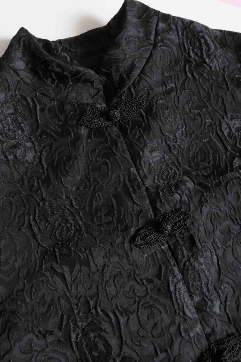 Saco negro de brocato con textura de rosas y alamares de pasamanería, mangas princesas y cuello mao, con bolsillos con solapa delanteros y largo a la cadera. 