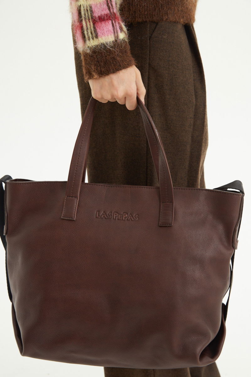 Cartera de cuero estilo shopper con logo grabado al frente, manijas cortas de cuero y correa larga de cinta mochilera.